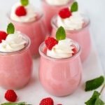 Pink mousse in glass cups topped with white cream and fresh raspberries.