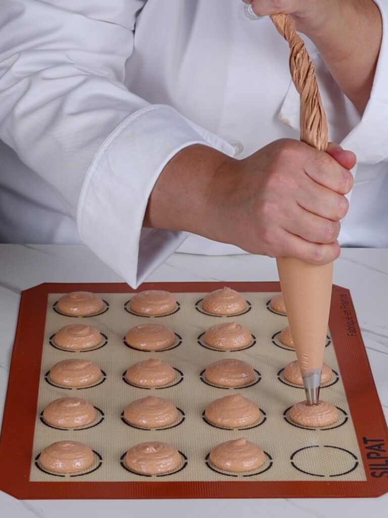 Hand is piping brown macaron shells.