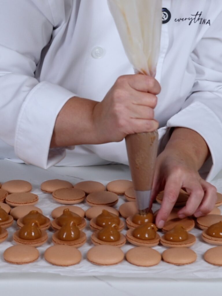 hand is piping salted caramel on a macaron shell.