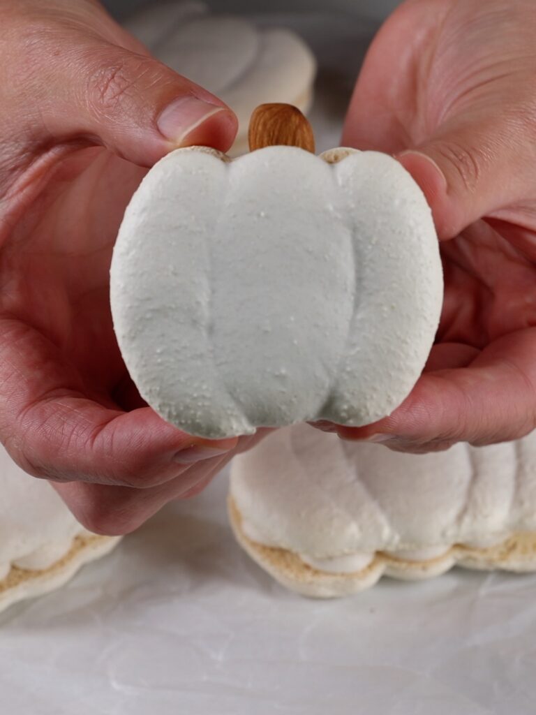 Hands are holding a pumpkin-shaped macaron.