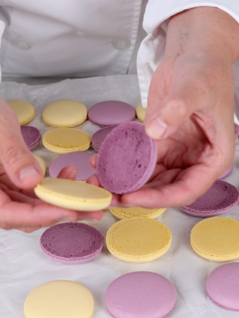 Hands holding yellow and purple macaron shells. 