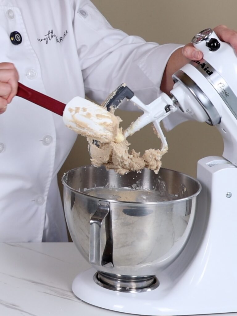 The hand is holding a spatula and a cleaning paddle attachment in the mixer.