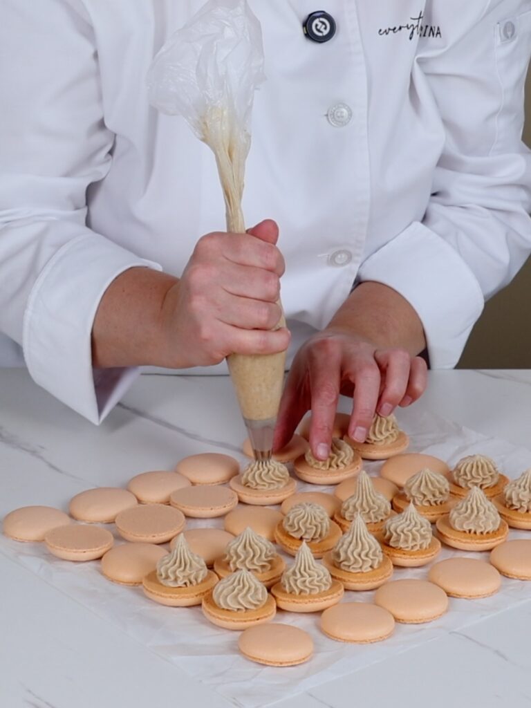 The hand is piping buttercream on the macaron shells.