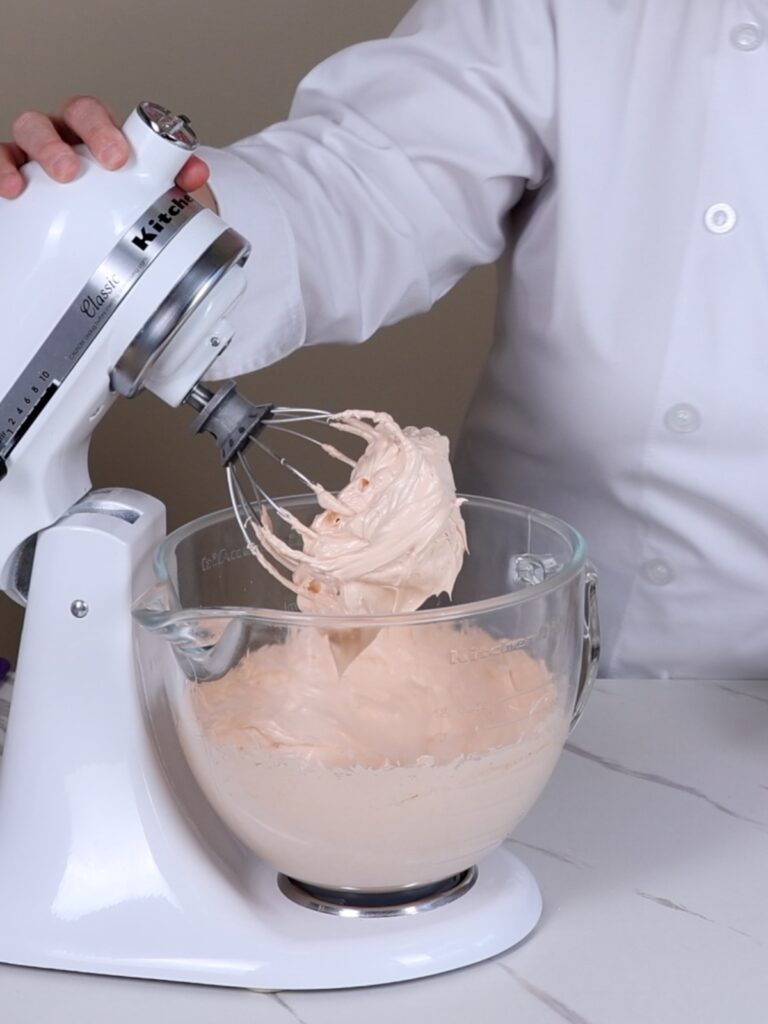 Orange French meringue in the mixing bowl of a stand mixer.