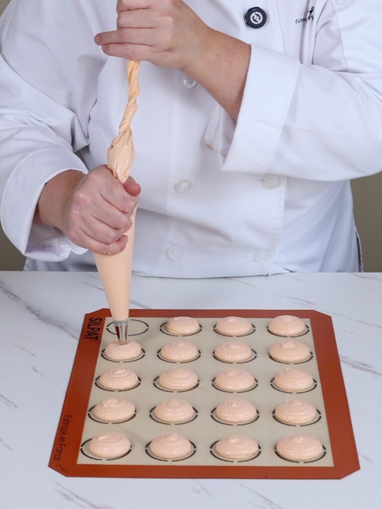 Hand is piping orange French macarons.