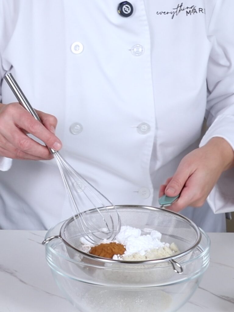 Hands are sifting dry ingredients into the mixing bowl.