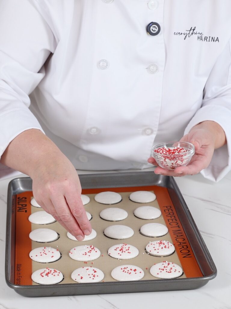 The hand is dusting sprinkles over white macaron shells.