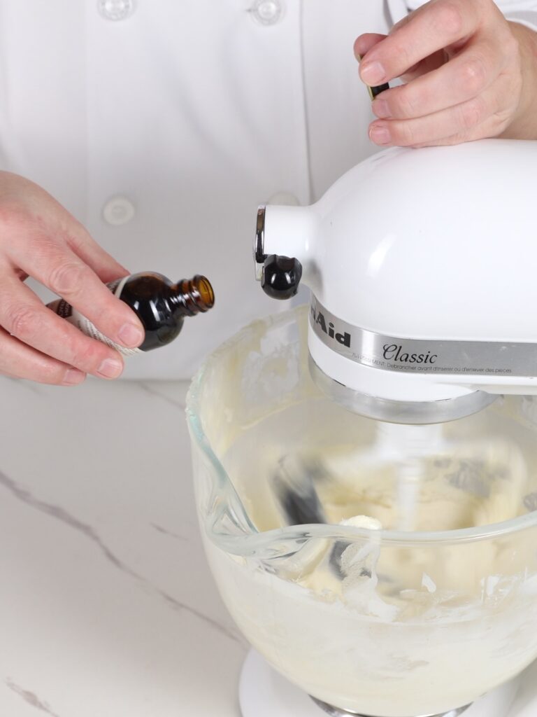 The hand is adding vanilla extract to the mixing bowl.