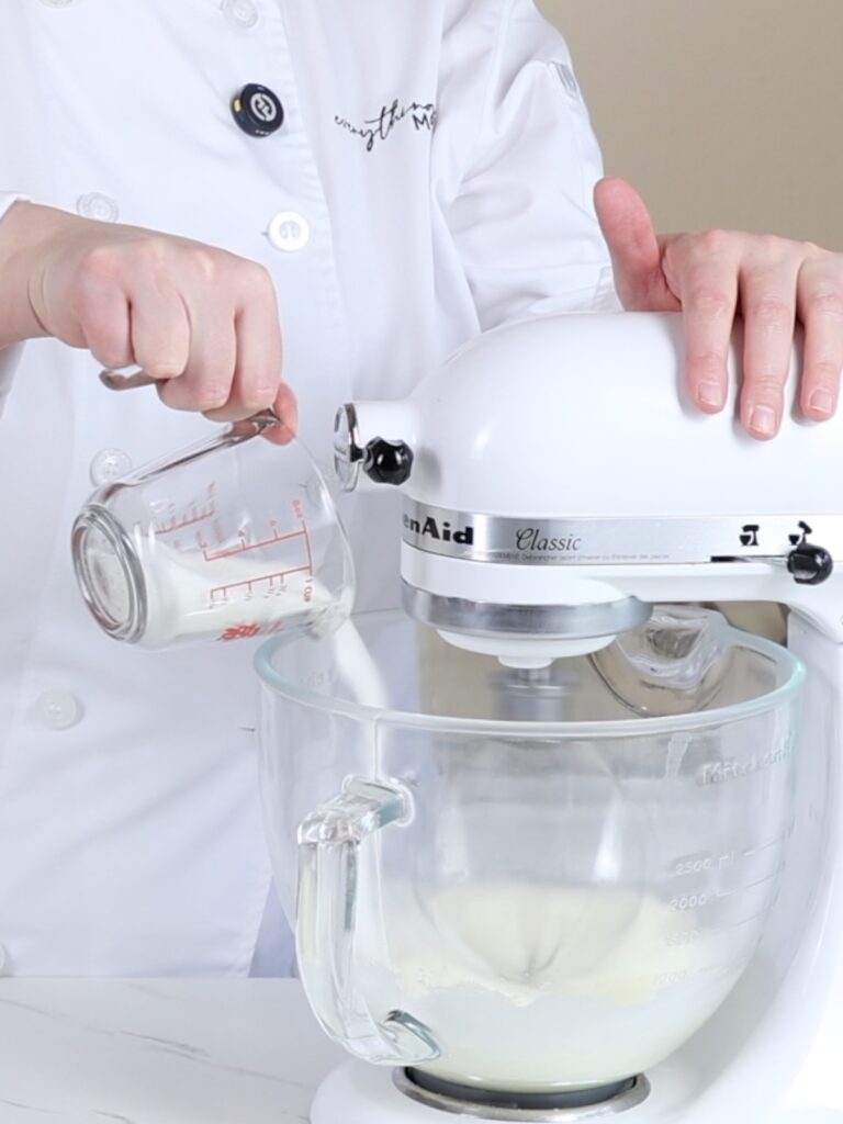 Hand adding sugar to the mixing bowl of the stand mixer. 